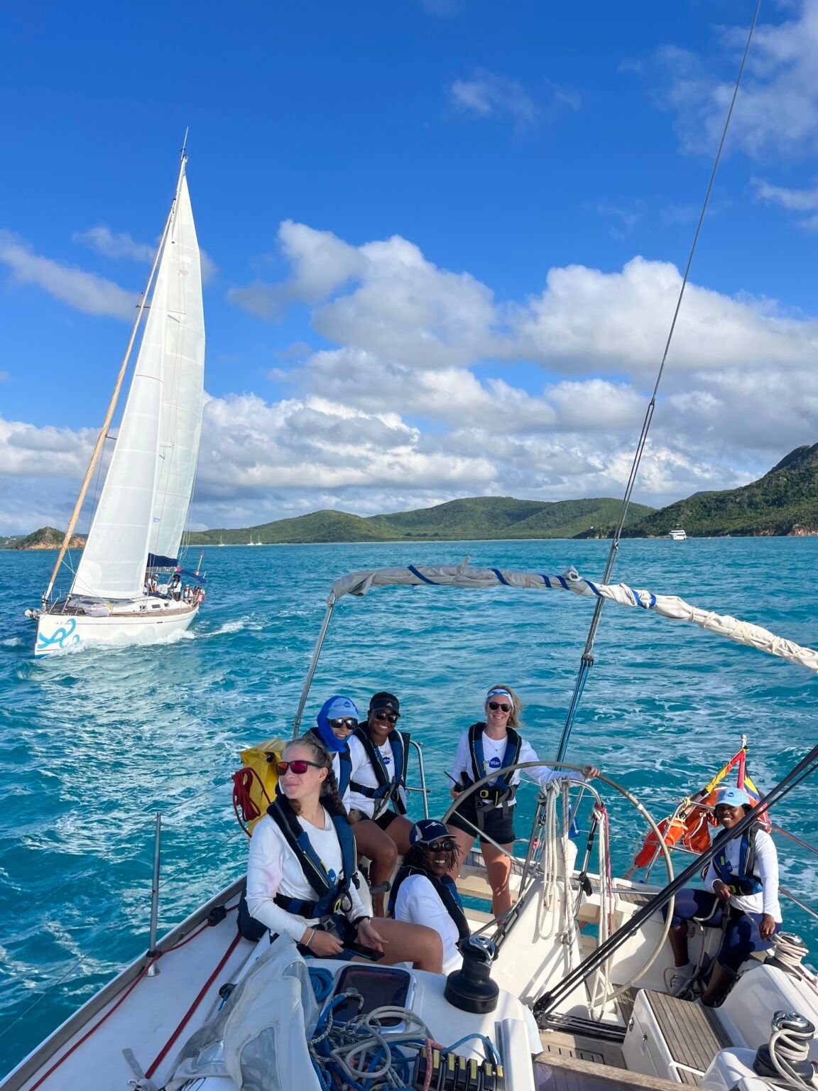 Six sailors in life jackets aboard a Second Star Sailing yacht, with another yacht on turquoise water behind them
